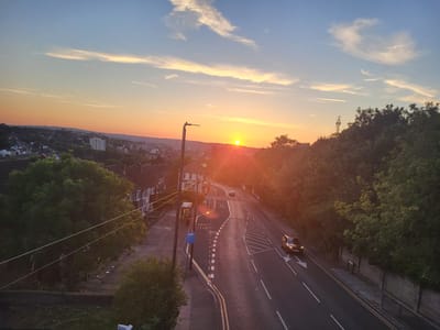 This is a high-angle, wide shot of a suburban street in Gillingham, United Kingdom, during a sunset. The sky is a gradient of blue and orange, with scattered clouds. The sun is setting in the distance, casting a warm glow over the scene.
In the foreground, there are power lines stretching across the frame. To the left, a row of residential houses with pitched roofs are visible. To the right, a road curves through the scene, with two cars driving on it. One car, a dark colored SUV, is in the right lane, and another car is further up the road. There is a bus stop with a shelter on the left side of the road. Trees line the right side of the road, and a hedge is visible at the edge of the road. In the distance, more houses and buildings can be seen, with a telecommunications tower on a hill.
