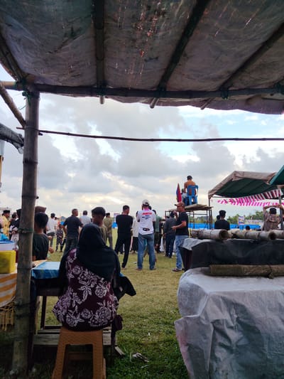 The image depicts an outdoor gathering in Cibrekbaroh, Indonesia, possibly a community event or market. In the foreground, a woman wearing a hijab and a patterned maroon and white dress sits on a plastic stool. She is seated at a table covered with a blue and white checkered cloth, partially obscured by a bamboo pole and a tarp-like structure overhead, constructed from bamboo poles and weathered tarpaulin. To the left, a yellow container and a bottle are visible on another table.
The middle ground shows a crowd of people standing on a grassy field under a cloudy sky. Many are casually dressed. Some individuals are engaged in conversation, while others appear to be observing something or waiting. A man in a white t-shirt with "PANITIA PIALA" and "ARI" text is visible. To the right, a raised wooden platform supports a person sitting on a blue plastic chair, facing away from the camera. A colorful flag is mounted behind this platform. Further to the right, a tent structure with a brown tarp and pink and white striped fabric is present. In the immediate foreground right, a table is covered with a light-colored tarp, with several lengths of bamboo resting on top. The overall scene suggests a lively, informal event in a rural or semi-rural setting.