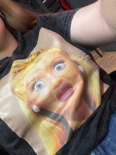 A close-up, high-angle shot shows the torso of a person wearing a black t-shirt with a printed image of a distorted, blonde woman's face. The print is large and takes up most of the frame. The person's right arm is visible, resting on the t-shirt. The background is out of focus, with hints of dark furniture and a wooden floor. This image was not taken in Alcalá De Henares, Spain.