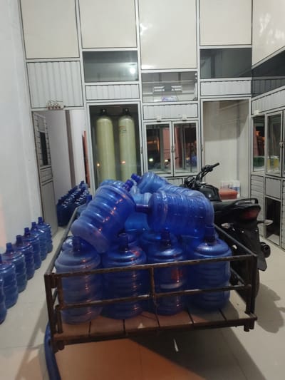 The image depicts an indoor storage area filled with numerous blue plastic water jugs. In the foreground, a cart loaded with these water jugs is prominent, with several jugs stacked on top. To the left, rows of blue water jugs are neatly arranged on the floor. In the background, there is a black motorcycle parked behind the cart. The environment appears to be a commercial space, possibly a warehouse or distribution center for bottled water, suggested by the large quantity of jugs and some shelving units with what appear to be water filtration tanks. The lighting is dim, casting shadows. The location is identified as Meurandehpaya, Indonesia. There are no people visible in the image.