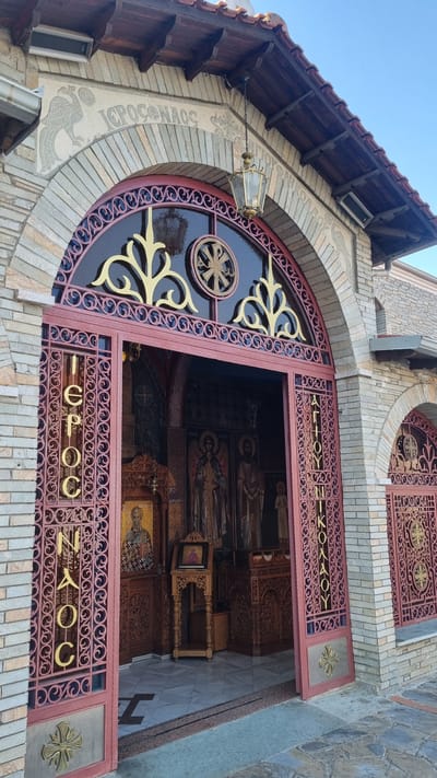 The image shows the entrance to a Greek Orthodox church in Litochoro, Greece. The facade is constructed of light-colored brick with stone accents. An arched entryway is framed by ornate, dark red metal gates with gold lettering and decorative elements. Above the entryway, the word "ΙΕΡΟΣ ΝΑΟΣ" (Hieros Naos), meaning "Holy Church," is carved into the stone arch. Inside the church, visible through the open gates, are religious icons and ornate wooden furnishings. A hanging lantern is suspended from the roof above the entrance. The ground in front of the church is paved with irregular stone slabs. The sky is clear and blue.
