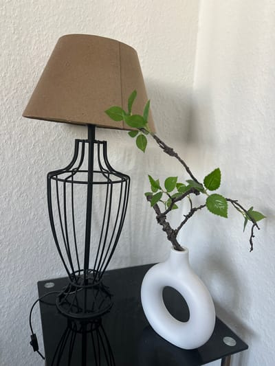 A bedside table in a room in Bielefeld, Germany, holds a black metal lamp with a brown lampshade. Next to the lamp is a white donut-shaped vase with a branch and green leaves. The table is made of black glass and has a metal frame. The wall behind the table is white and textured. A power cord for the lamp is visible on the table.