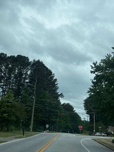 The image depicts a street in Mountain Park, United States of America on an overcast day. The road is paved with yellow lines indicating lanes. A "STOP" sign is visible on the right side of the road, accompanied by a red light illuminated above. Dense foliage of trees lines both sides of the street. Utility poles with numerous power lines crisscross the view above the trees.