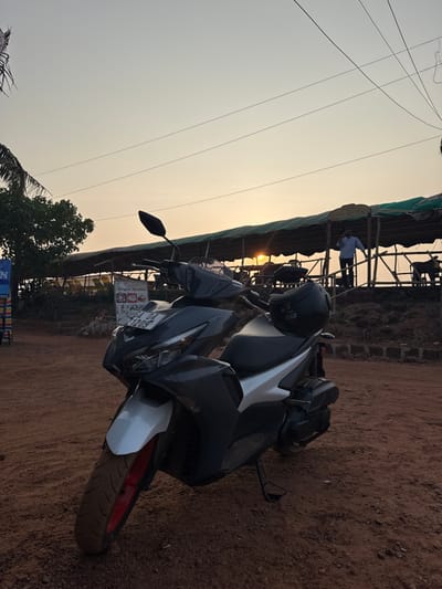 A gray and white Yamaha Aerox scooter, license plate GA 04 2120S, is parked on reddish-brown dirt in Bhagatwadi, India, at sunset. A black helmet rests on the scooter's rear seat, and its front wheel exhibits a red rim. In the background, an elevated open-air structure with a green tarp roof and wooden supports is present. A male individual in a light-colored shirt stands on the structure's upper level, silhouetted against the setting sun, holding an object near his ear. Two dark-colored animals are visible beneath the structure. Overhead utility lines are present against the warm, clear sky.