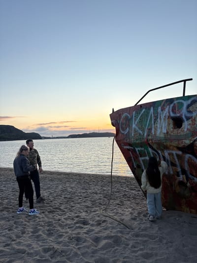 This image depicts a sunset scene on a sandy beach in Teriberka, Russia, adjacent to a body of water. In the foreground, a partially submerged shipwreck, heavily vandalized with graffiti, dominates the right side of the frame. A young girl with long dark hair, wearing a light-colored jacket and blue pants, stands with her back to the camera, touching the graffiti on the shipwreck. To the left, a man and a woman stand on the sand, looking out towards the water and distant hills. The man is wearing a camouflage jacket, and the woman is wearing a dark jacket and black leggings. The sky above is a gradient of blue and pale orange, indicating the setting sun, with clouds on the horizon. The water is calm, reflecting the sky's colors. The sandy beach is textured with ripples and footprints.