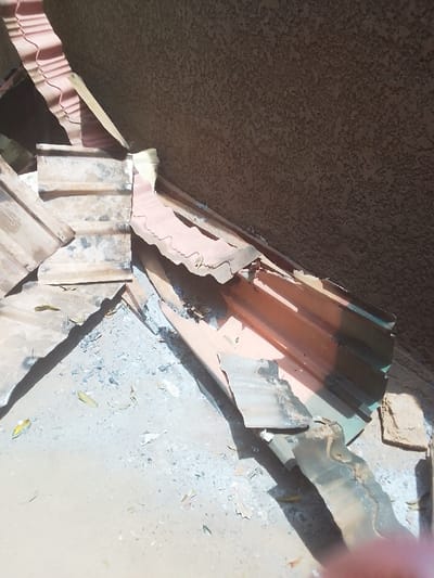 The image shows a pile of broken corrugated metal roofing sheets lying on a concrete surface in front of a textured wall. The debris is scattered around a patch of grey ash. There are no people or animals in the image. The location is a residential area in Kwal, Nigeria, based on the provided coordinates.
