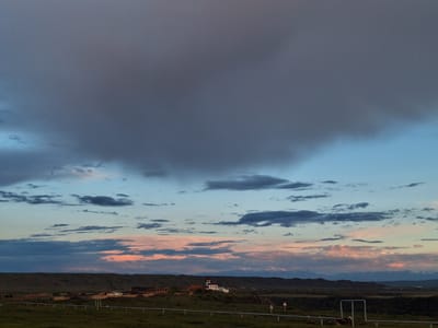 The image presents a wide-angle view of the city of Anipemza, Armenia, taken during a sunset with dramatic cloud formations. The lower third of the frame is dominated by a green field, crossed by what appear to be white metal guardrails. In the middle ground are clustered buildings, likely part of the city itself, with a white, two-story building with a red roof standing out prominently on a small rise. In the distance, low hills meet the horizon, behind which a faint line of snow-capped mountains is visible. The sky is a mix of blues, pinks, and dark grays, indicative of sunset conditions. The cloud cover is dense, with large, dark clouds occupying the upper portion of the image.
