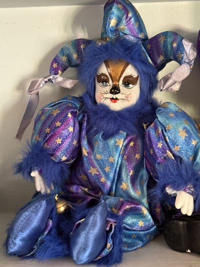 The image shows a close-up, head-on view of a doll dressed in a jester-like costume. The doll has a porcelain face with painted features resembling a cat or fox, with blue eyes, a black nose, and red lips. It wears a blue and purple striped jester hat adorned with gold stars, and a blue furry collar. The doll's costume is also blue and purple with gold stars, featuring puffy sleeves trimmed with blue fur. The doll is seated, with its blue satin legs crossed and a small gold bell attached to its waist. The background is a plain, light-colored wall, suggesting the doll is displayed indoors. The image was taken in the city of Graham, United States of America.