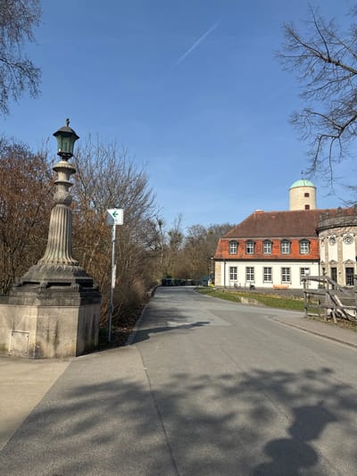 This image depicts a daytime scene in Bad Nauheim, Germany, under a clear blue sky with a visible contrail. The foreground features an ornate, weathered stone streetlamp with a dark lantern fixture, positioned on the left side of an asphalt path. To the right of the lamp, a white rectangular sign with a green left-pointing arrow and a bicycle symbol is affixed to a pole.
The path curves to the right and extends into the midground. On the left side of the path, bare deciduous trees and shrubs line the edge, casting shadows across the asphalt. In the distance along this left edge, several dark-colored refuse bins are visible.
To the right of the path, a two-story building with a red-tiled hip roof and white facade occupies the midground. The building features multiple evenly spaced windows with white frames and closed shutters on its ground floor. An attached section on the right exhibits Baroque-style architectural ornamentation with round windows. A light green-domed tower with small rectangular windows rises from the building's right side. Several bicycles are parked in front of the building, and a low wooden fence or structure is visible on the far right. No individuals are present in the scene.