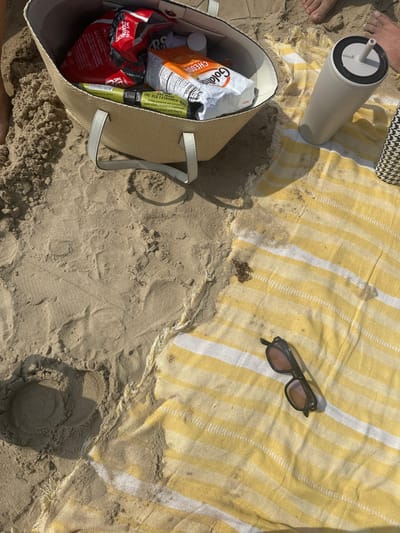 An overhead view of a sandy beach scene in South Padre Island, United States of America, under direct sunlight. A light-colored, woven tote bag with two handles rests on the sand in the upper left, containing a red crumpled bag (possibly chips), a white bag of Goldfish crackers, and a green tube labeled "The Old Fashioned." Bare feet are partially visible in the upper left and upper right corners. A matte beige tumbler with a black lid and straw stands upright on a yellow and white horizontally striped beach towel. The towel, positioned in the lower right, shows signs of sand, moisture, and scattered dark debris. A pair of dark-framed sunglasses with tinted lenses lies on the towel in the lower right. The sand surface exhibits footprints and impressions.