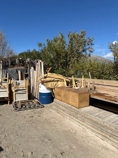 This outdoor image captures a storage area or yard under a clear, bright blue sky in Anatoliki Attiki, Greece. The scene is dominated by a variety of stored materials and natural vegetation.

On the left side of the frame, dense green foliage from large bushes or trees, with slender, olive-like leaves, fills the mid-ground, extending towards the sky. The bright sunlight indicates daytime and clear weather.

The right side and foreground are filled with various stored items. A large quantity of wooden planks and lumber is stacked horizontally, forming a wall-like structure that runs from the mid-ground towards the background on the right. In front of these stacks, two light brown wooden crates are stacked vertically, with the word "CORONA" clearly visible on their sides. Next to these crates, a blue barrel with a white top section is partially covered by a tan tarp or fabric.

Further into the foreground and towards the bottom, more miscellaneous wooden materials are piled, including a visible wooden pallet on the dirt and gravel ground. Stacked behind this pallet are items that appear to be chairs or table tops with white woven patterns, resting on wooden frames or smaller pallets. The ground is a mix of dirt and small stones. No people are visible, and no specific activity is taking place, suggesting a static storage or staging area.
