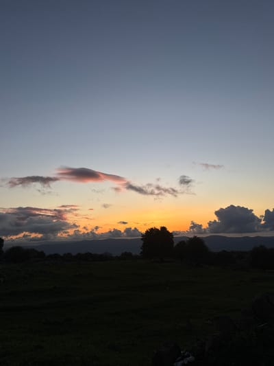 The image depicts a sunset over a rural landscape near Mas'ade, Israel. The sky transitions from a light blue on the left to an orange and yellow gradient near the horizon. Several dark clouds are visible near the horizon and towards the left side of the image, with some showing hints of pink coloring from the sunset. The lower right portion of the image shows a dark field or meadow, with a line of trees silhouetted against the colorful sky. In the distance, a subtle outline of hills or mountains can be discerned through the clouds. No people or signs of human activity are readily discernible beyond the cultivated landscape.