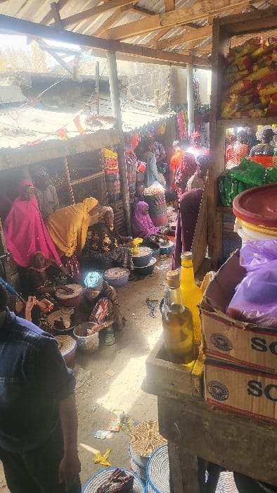 This image depicts a bustling market scene located in Fagge D1, Nigeria. The environment is a semi-covered structure with a corrugated metal roof supported by wooden beams, allowing direct sunlight to penetrate and cast strong shadows. The ground is uneven, appearing to be dirt or rough concrete, with scattered debris including paper and plastic.
Numerous individuals are present, predominantly women, many wearing traditional head coverings and dresses in various colors such as pink, yellow, purple, and patterned brown. Most of these women are seated directly on the ground or on low stools, actively engaged in selling or preparing goods. They are positioned around large bowls and containers of varying colors (white, blue, black, red) which hold dry goods, possibly grains or powders. Several stacks of patterned woven baskets, both blue and brown, are visible on the ground as display or storage.
In the immediate foreground, to the right, a wooden stall displays two glass bottles containing yellow liquid (likely oil), purple plastic bags, and cardboard boxes marked "SCO." To the left foreground, a person wearing a blue plaid shirt and dark pants is partially visible, facing away from the camera. In the midground and background, additional market activity continues with more people standing and seated, and goods displayed on shelves, including stacks of yellow and red packaged items and green fabrics. The background reveals unfinished or damaged concrete walls of adjacent structures, and colorful decorative bunting strung across the market area.