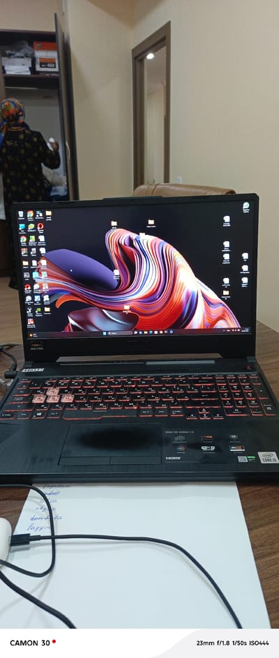 This is a medium-shot, indoor, eye-level photograph taken in the city of Türkmenbaşy, Turkmenistan. The primary subject is a black ASUS TUF Gaming laptop placed on a wooden desk. The laptop screen displays a colorful abstract wallpaper with flowing shapes and is turned on, showing various application icons on the desktop. The keyboard has red backlighting and features Cyrillic characters. To the left of the laptop, a person wearing a headscarf and a patterned garment is partially visible, appearing to be interacting with something inside an open wardrobe. In the foreground, a white sheet of paper with handwritten text in a blue pen is visible, along with a black USB-C cable and a white earbud. The background shows a wardrobe with shelves and a doorway with a mirror. The overall lighting suggests an indoor environment, possibly a room in a residence or office.