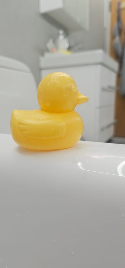 A yellow rubber duck is positioned on the edge of a white bathtub. The duck is facing right, with its beak slightly raised. The surface of the bathtub is wet, with small droplets of water visible. In the background, a blurred image of a bathroom sink and cabinets can be seen, with a faucet and some toiletries on the counter. The overall scene suggests a typical bathroom setting, possibly after a bath or shower. The image does not contain any elements or context that would allow for its localization in Tynset, Norway.