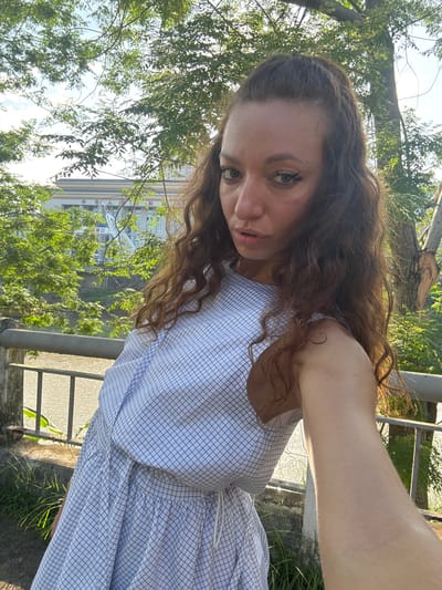 A woman is taking a selfie outdoors, positioned next to a metal railing. She is wearing a light blue and white checkered dress. Her hair is long and wavy. The background shows green foliage, a glimpse of a building, and part of a city scene, suggesting the photograph was taken in a semi-urban environment. The building visible in the distance features Chinese characters, which corroborates the location in Lao Cai Ward, Vietnam, given its proximity to the Chinese border. The lighting is bright, suggesting it is daytime.