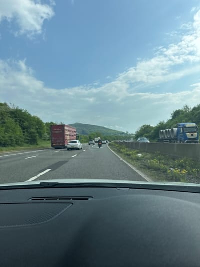 The image depicts a highway scene, likely in the vicinity of Woolvers Hill, United Kingdom, based on the surrounding landscape and road infrastructure typical of the region. The view is captured from inside a vehicle, specifically from the passenger side. A line of traffic is visible ahead, including a large truck with a red cargo container and several other cars and trucks, a motorcycle in front of a white car. The road is flanked by green vegetation and trees, with rolling hills in the background. The sky is partially cloudy with patches of blue. The dashboard of the vehicle is visible in the foreground.