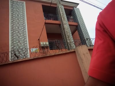A low-angle, medium shot captures a section of a coral-colored, two-story building in Nanka, Nigeria. A vertical concrete column, adorned with a patterned grey tile design, divides the facade. The building features balconies with black metal railings and a single window visible on the upper floor. A rough, coral-colored wall with barbed wire on top runs across the foreground. To the right, a person wearing a red shirt is partially visible, their arm and shoulder in frame. The sky is overcast. The image captures architectural details, security measures, and a hint of human presence within an urban environment.