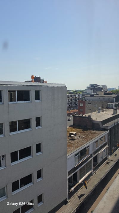 This media file shows an aerial view of a cityscape, likely taken from a high vantage point such as a drone or a tall building. The scene is dominated by buildings of varying heights and architectural styles, with a clear blue sky occupying the upper portion of the frame. The orientation of the image is rotated, presenting the cityscape as if viewed from the side.
In the foreground, a modern building with a grey facade and multiple large, rectangular windows is prominent. Further into the background, a mix of residential and commercial structures can be observed. Some buildings have flat roofs, while others appear to have pitched roofs covered in what looks like vegetation or moss. Narrow streets or alleys are visible between some of the buildings. One building has a distinctly red brick facade.
The weather appears to be clear and sunny, suggesting daytime. The time of day is likely midday or afternoon, given the angle of the sunlight casting shadows on the buildings and the overall brightness. The presence of a yellow and blue flag on a roof and the architectural style of the buildings hint at a European setting, specifically Vlaardingen, Netherlands, as per the context.
There are no people clearly visible in the image. Some objects like ventilation pipes or rooftop structures are scattered across the rooftops. A subtle watermark in the lower right corner reads "Galaxy S26 Ultra," indicating the device used to capture the image. The overall atmosphere is one of an urban environment on a bright, clear day.