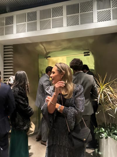 A group of people appear to be inside or entering a venue. A blonde woman in a shimmery black dress is the focal point. A man in a gray suit walks behind her. Other attendees are partially visible, including a woman in a green dress and black fur stole. Plants decorate the background. An interesting architectural design appears on the left wall with patterns on square shapes.