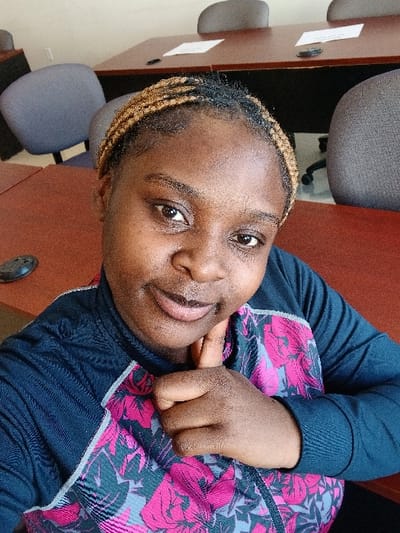 A woman with braided hair is indoors, wearing a dark blue jacket with pink floral designs. She's looking at the camera with a slight smile, her right hand near her face. The background features a reddish-brown surface, possibly a table, with chairs. A black, round object is visible on the table. The scene is brightly lit.