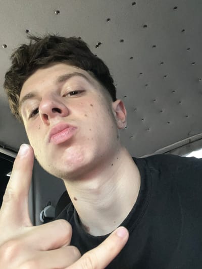 The image is a close-up, low-angle selfie of a young Caucasian male. He has short, dark, messy hair and is wearing a black t-shirt. He is making a duck face and has his right hand raised, with his index finger and thumb forming a "gun" gesture. His left hand is also visible, with his index and middle fingers extended in a "peace" sign. The background appears to be the interior of a vehicle, with a textured ceiling and what looks like a window on the right side. There are no discernible elements that would indicate the location within Leshtak, Bulgaria.