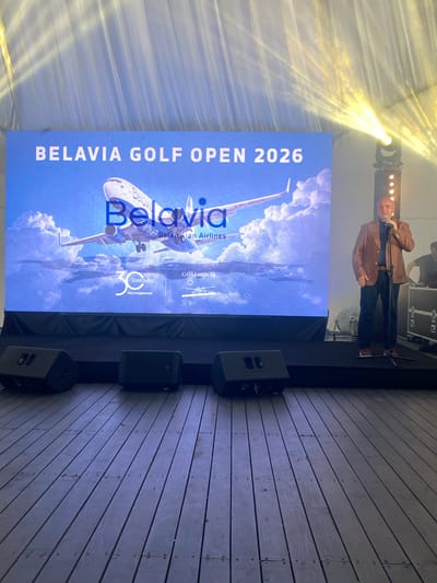 The image depicts an indoor event space with a large LED screen displaying "BELAVIA GOLF OPEN 2026" and an image of a Belavia airplane against a cloudy sky. Below the main text, a logo with "30" and "Belavia" is visible, along with the text "МЫ СОЕДИНЯЕМ" which translates to "WE CONNECT." To the right of the screen, a man in a brown blazer and dark pants stands on a stage, holding a microphone and speaking. He is illuminated by a spotlight. The stage has black carpeting, and black speakers are placed on the wooden floor in front of it. In the background, other equipment and personnel are faintly visible. The overall scene suggests a promotional event or a conference. The location of Kalodziešča, Belarus is implied by the presence of Belavia, a Belarusian airline, and the Belarusian text on the screen.