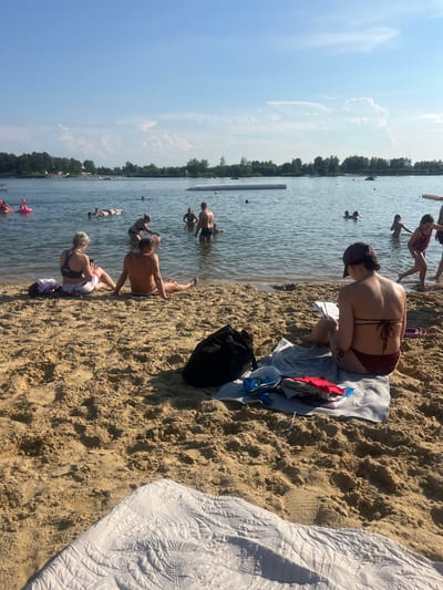 This is a photograph taken at the Cholerzyn Lake in Cholerzyn, Poland. The scene depicts a sunny day at a beach resort. In the foreground, on a sandy beach, two individuals are sitting. One, wearing a maroon bikini and a cap, is looking down at a book or tablet. Next to her, a black backpack and folded clothing are visible on a gray blanket. Further back, a couple sits on the sand, with the woman facing away from the camera and the man facing sideways. The lake is populated with people swimming and floating on inflatable devices. The background shows a treeline with buildings and a clear blue sky with scattered clouds.