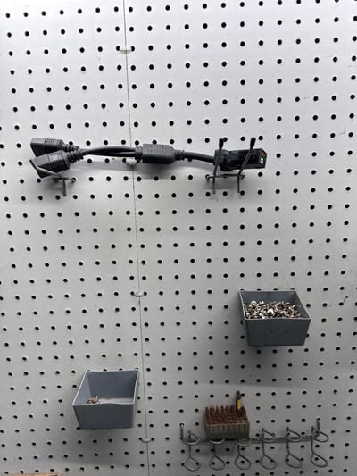 This image depicts a pegboard wall in what appears to be a workshop or storage area, likely located in Indiantown, United States of America. The pegboard is white and features a uniform pattern of circular holes.

Mounted on the pegboard in the upper section are two black electrical cables connected to a power strip or adapter. The cables are secured to the wall with metal hooks. Below the cables, to the right, is a grey plastic bin filled with small metal fasteners, possibly screws or bolts.

Further down on the pegboard, to the left, is another grey plastic bin, this one containing a smaller quantity of similar metal fasteners. Below this bin is a rack made of wire that holds a set of drill bits or screwdriver bits in a grey plastic holder. Several other wire loops, possibly for hanging tools, are also visible on this rack.

The overall scene is one of organized storage for electrical components and hardware. There are no people visible in the image.