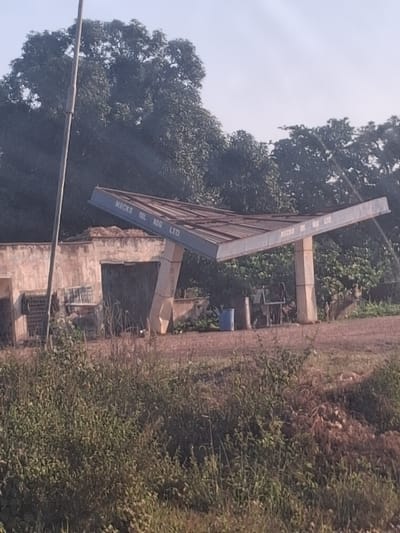 This image displays an unpaved outdoor scene in Mutum Biyu, Nigeria, centered on a disused fuel station canopy structure. The canopy, colored blue-grey, bears the white inscription "MICKS OIL NIG LTD" on its visible underside and is supported by two prominent, light-colored concrete pillars. To the left of the canopy, a dilapidated light-brown concrete wall structure, featuring an opening and stacked metal grates, is visible. The background consists of dense green trees and foliage against a hazy, bright sky, with a tall, dark vertical pole positioned on the far left. The ground immediately beneath and around the canopy is reddish-brown dirt or gravel. Near the base of the right concrete pillar, a blue plastic barrel and indistinct dark objects, possibly furniture, are present. The immediate foreground is dominated by abundant, unkempt green and brown grasses and weeds, with a small mound of earth or debris visible on the right. No individuals are present, and no actions or interactions are observed.