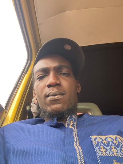 The image shows a man with a beard, wearing a black baseball cap and a blue patterned shirt. He is inside a vehicle with yellow interior and beige seats. The setting could be in a car or taxi in Ijaiye, Nigeria. The image is rotated, showing the man's face at an angle.