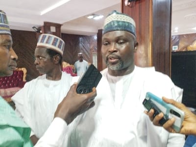 The image shows three men standing in an indoor setting, likely a government office or public building, in Kaltungo, Nigeria, at coordinates 9.816, 11.315. The man in the center is wearing a white long-sleeved shirt, a blue and white cap, and a short grey beard. He is being interviewed by another man holding a black digital recorder and microphone. The man on the left is wearing a white long-sleeved shirt, a colorful patterned cap, and is looking to the left of the frame. He is partially obscured by the man in the center. The man on the right is wearing a blue shirt and is holding a blue and black cell phone. He is only partially visible in the frame. There are wood-paneled walls and a red plush chair behind the men.  The environment appears to be a formal setting. The men are interacting in a professional manner, with the man in the center being the subject of the interview. The room has wood-paneled walls and a red plush chair, and a black electrical outlet is visible on the right wall. The lighting appears to be bright, and the image is well-lit.