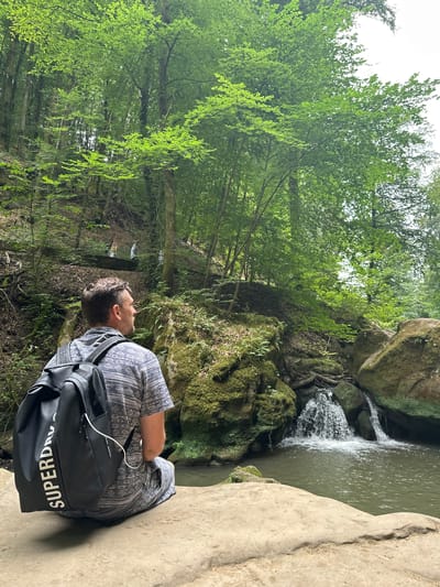 In Waldbillig, Luxembourg, a man with short brown hair sits facing away from the camera on a large, light-colored rock. He is wearing a short-sleeved shirt with a grey, blue, and white patterned design and matching shorts. A large, black backpack with white lettering that reads "SUPERDRY" is on his back, with a white cord extending from it. He is looking towards a wooded area where a small waterfall cascades into a pool of water. The waterfall is surrounded by moss-covered rocks and lush green trees. In the background, on a higher elevation, three people can be seen walking. The environment is a natural landscape characterized by dense foliage and rocky terrain.