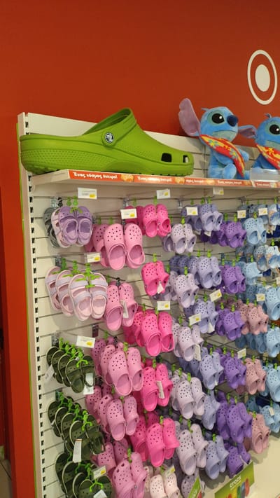This image depicts a retail display of Crocs footwear in a store, likely a shoe store or a department store. The display features several rows of brightly colored Crocs, primarily in shades of pink, purple, light blue, and green. The shoes are hung on a white pegboard system. Above the Crocs, a large, lime green Croc shoe is displayed horizontally. To the right of the large green shoe, two plush toys resembling characters from the Disney movie Lilo & Stitch are visible. A circular logo with concentric white rings is visible on the orange wall behind the display. The store appears to be well-lit and organized, with rows of merchandise extending into the background. The presence of Greek text on signage suggests the location is in Greece. The store is identified as being located in the city of Pylaia Municipal Unit, Greece.