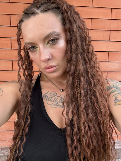 This is a vertical, close-up, selfie-style photograph of a person with long, wavy, auburn hair that is braided in some sections. The person's face is turned towards the camera, with their eyes open and looking slightly upward. They are wearing a black tank top and have several tattoos visible on their chest and arm, including script lettering on the chest. A silver chain with a spherical pendant is visible around their neck. The person is also wearing a silver nose ring. Their makeup includes green eyeshadow.
The background consists of a wall made of reddish-brown bricks laid in a horizontal pattern. The lighting suggests it is daytime, possibly outdoors due to the natural light. The overall mood appears casual and personal. No specific activity or event is depicted beyond posing for the photograph. The location context provided is Grushevskaya, Russia, but there are no specific visual cues in the image to confirm this location.