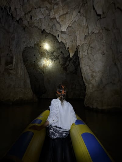 This image depicts a person navigating a water-filled cave in the city of Phang-nga, Thailand. The scene is illuminated by a headlamp, casting bright light on the cave's limestone ceiling and upper walls while leaving the majority of the environment in darkness.

A single individual, seen from behind, occupies the foreground. They possess medium-length dark hair, tied back with an orange hair tie. An orange headlamp strap, bearing visible text "HEADLIGHT" and "STARTS," is worn on their head. They are dressed in a white long-sleeved shirt and patterned dark and white shorts or trousers. Their right hand rests on the yellow side of an inflatable boat, which features prominent yellow and blue sections and occupies the lower portion of the frame.

The cave interior reveals rugged, light-colored rock formations on the ceiling and walls, with some visible stalactites. Numerous bats are observed hanging from the cave ceiling and upper walls, particularly within the illuminated areas. The cave floor is submerged in dark water, reflecting minimal light. The overall atmosphere is dim, with the headlamp providing the primary light source for observing the cave's natural features and inhabitants.
