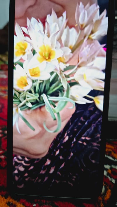 A close-up, vertically oriented, low-angle, outdoor photograph shows a person's hand holding a bouquet of small, white and yellow flowers with green foliage. The flowers are clustered together and appear delicate. The person's hand is visible from the wrist down, cupping the base of the bouquet. The background consists of a dark, patterned fabric and a red and orange patterned rug, suggestive of an indoor setting with decorative textiles. The image is captured on a mobile device, indicated by the black border framing the scene. The photograph does not provide any specific information about the city of Kaka, Turkmenistan, or any other geographical location.