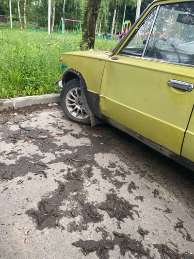 The image depicts the side of a yellow vintage car parked on a paved area. The car shows signs of wear, including rust around the wheel arch and minor scratches on the door. The ground is wet and strewn with debris, possibly leaves and twigs, suggesting recent rain or that the area is unkempt. In the background, a grassy area with trees and a playground with swings are visible, indicating an urban or semi-urban setting. The presence of a playground suggests the location could be a park, residential area, or schoolyard in Khotkovo, Russia. There are no people visible in the image, and no specific actions are being depicted. The overall scene is static and focuses on the car and its immediate surroundings.