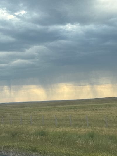 An overcast sky looms over a vast, open field of green grass in Төрт Құдық ауылдық округі, Kazakhstan. Several columns of rain are visible falling from the clouds, creating shafts of light in the sky. A wire fence with wooden posts runs horizontally across the middle of the frame, separating the foreground from the distant landscape. The terrain appears mostly flat, with subtle undulations. The overall atmosphere is one of a stormy, rural landscape.