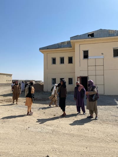 The image depicts an outdoor scene in Bar Mel, Afghanistan, under a clear, cloudless blue sky. The ground is unpaved, consisting of dry, light brown dirt and gravel, with various debris scattered across it.
In the mid-ground and background, multiple light-colored buildings are visible. The most prominent structure on the right is a two-story, light beige building with a damaged or incomplete corrugated metal roof, showing exposed structural elements. This building features multiple rectangular windows, some appearing vacant or without glass panes. An open doorway is present on the ground floor, and a grid-like metal frame is attached to the wall adjacent to it. In the distance to the left, additional similar-looking buildings are visible, suggesting a larger complex. A mound of earth or construction debris is present in the mid-ground between the groups of people.
Approximately ten to twelve adult males are present, dressed in traditional Afghan clothing, including shalwar kameez, turbans, and vests. In the right foreground, five men are standing. One man in a light brown shalwar kameez and a dark vest, facing left, has a rifle or long gun slung across his back and carries a small backpack. To his right, a man in a black shalwar kameez with a patterned vest and a white head covering adjusts his headwear. Further right, a man in a dark purple shalwar kameez with a reddish-brown scarf around his neck stands, looking towards the left. Partially visible on the far right is another man in dark purple attire. In the mid-ground and background to the left, several other men are walking from left to right, also in traditional dress. Strong shadows from the individuals are cast on the ground, indicating bright sunlight.