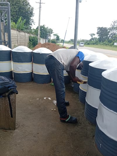 A man is meticulously applying masking tape to the center bands of several large cylindrical containers, presumably in Igrita, Nigeria. The containers are gray with white bands, and the tops are covered in a white substance. The man, dressed in a light-colored shirt, jeans, and clogs, is working outside near a road and a wall. A toolbox and a bag sit nearby.