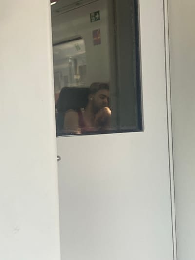 The image depicts a person with blonde hair, wearing a red tank top, sitting inside a train carriage. The person is looking down and appears to be resting their chin on their hand, possibly listening to something through white earbuds. The reflection in the window shows the interior of the train, including a seat and some signage. The exterior of the train carriage is white. The setting is likely within the city of Montcada i Reixac, Spain, as indicated by the available information.