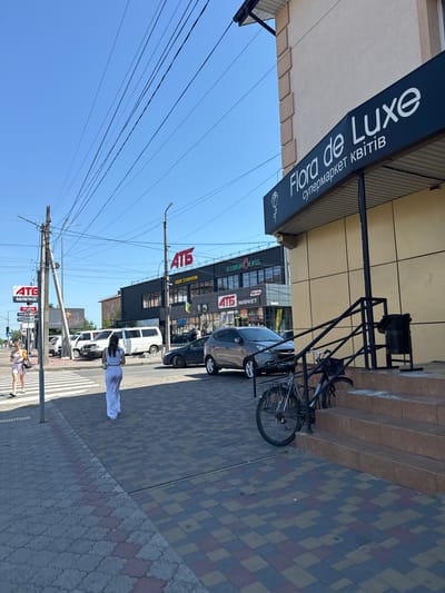 This media file depicts a street scene in Sarny, Ukraine, under a clear blue sky, suggesting a sunny day. The camera is positioned to capture a perspective looking upwards and slightly down the street.
In the foreground, a woman in a white outfit is walking across a pedestrian crossing. Further down the street, two other individuals, likely women, are also visible, one of whom appears to be walking a dog on a leash. A parked bicycle is attached to a railing near a building entrance. Several cars are parked along the street, including a white van and a darker sedan. A bicycle is also parked in front of a building with a dark facade.
The buildings visible include a store with a prominent sign reading "Flora de Luxe" which also states "супермаркет квітів" (flower supermarket). Adjacent to this is another establishment with signage that includes "ECONOMOLASS" and "ATG MARKET" with operating hours "00-24". Another sign reads "ATG" and "ОДЯГ З ЄВРОПИ" (clothing from Europe). Traffic signs are also visible, including one indicating a speed limit of 40 and another for a distance of 100 meters. A network of overhead power lines dominates the upper portion of the image. The street and sidewalks are paved with patterned bricks. The overall atmosphere appears to be a typical urban daytime scene.