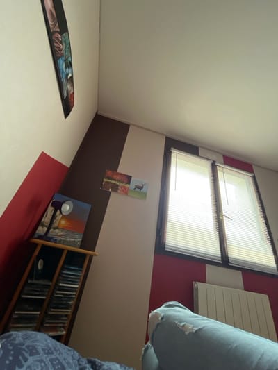 The image depicts an interior room in Ancenis, France, viewed from a low angle. The walls feature a geometric paint scheme consisting of white, red, and dark brown sections. On the right wall, a double window with a dark frame is fitted with white horizontal blinds, through which bright daylight is visible. A white radiator is mounted directly below this window.
On the left side of the frame, a multi-tiered wooden shelving unit is present, filled with numerous compact discs or digital video discs. The top shelf of this unit holds a purple-hued desk lamp with a white bulb and a framed picture depicting a sunset over water. Above the shelving unit, a long, narrow artwork composed of multiple segmented images hangs on the wall. Further to the right, on a white wall section, two smaller framed pictures are displayed: one depicting autumnal trees and the other a deer in a field.
In the immediate foreground, the lower half of a person's legs is visible, clad in light blue ripped jeans, specifically showing a tear on one knee. A dark blue patterned blanket or duvet is partially visible in the bottom left corner, suggesting the person is in a reclining position.