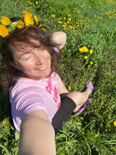A woman is lying in a field of grass and dandelions. She is wearing a pink t-shirt and black shorts. She is wearing a crown of dandelions on her head, and purple sandals on her feet. She is smiling and looking at the camera. The location is not in the city of Узяр, Russia.