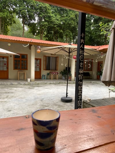 The image depicts an outdoor courtyard scene, likely a cafe or restaurant, located in Dujiangyan City, China. A wooden table occupies the foreground, on which a patterned ceramic cup rests, partially obscuring the view of the courtyard. The courtyard itself is paved with concrete and features a building with several wooden doors and windows. Umbrellas are set up, suggesting an outdoor dining or seating area. Decorative elements like string lights with stars and crescent moons, as well as paper lanterns, are visible, indicating a festive or decorative atmosphere, possibly for an evening setting or event. Lush green trees surround the courtyard, contributing to a natural and serene environment. A black metal pole with Chinese calligraphy written on it stands prominently in the courtyard, adding a cultural element to the scene. The overall mood appears calm and inviting. The time of day is likely late afternoon or evening, given the presence of string lights, and the weather appears to be overcast or twilight.