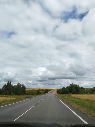The image shows a view from a car driving on a paved road through a rural landscape in Medvezh'ya Polyana, Russia. The road curves to the right in the distance. On either side of the road are fields of golden crops and green trees. The sky is filled with large, gray and white clouds, with patches of blue sky visible. The scene appears to be during the daytime. The bottom of the image shows a portion of the car's dashboard and windshield.