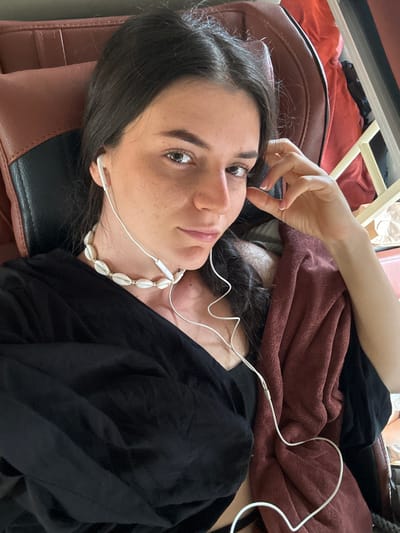 A woman is lying in a brown leather chair. She is wearing a black shirt, a seashell necklace, and white wired earbuds. A brown blanket is draped over her. Her hand is near her face. The location is potentially Đại Ninh, Vietnam, but there is no information in the image to verify this.