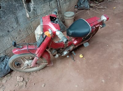 A red moped-style motorcycle is parked on a dirt ground surface. The motorcycle appears weathered and possibly in need of repair. A black vinyl seat is mounted on top of the motorcycle's frame. A yellow fruit is on the ground. The motorcycle is positioned close to a concrete wall. The location is Orji, Nigeria.