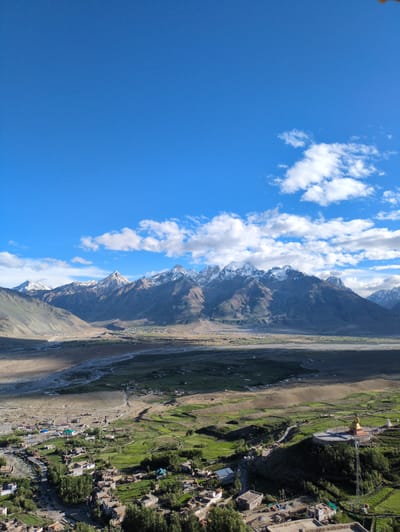 This is a high-angle, vertical, full shot of a mountainous landscape with a village in the foreground. The sky is a bright blue with scattered white clouds. In the middle ground, a range of snow-capped mountains dominates the horizon. Below the mountains, a valley unfolds with a riverbed and a small village nestled within it. The village consists of numerous small buildings with brown roofs, surrounded by patches of green vegetation and trees. In the foreground, on a hill to the right, a large golden Buddha statue stands prominently. A telecommunications tower is also visible near the statue. The overall scene depicts a remote and serene mountainous region in Yulang, India.